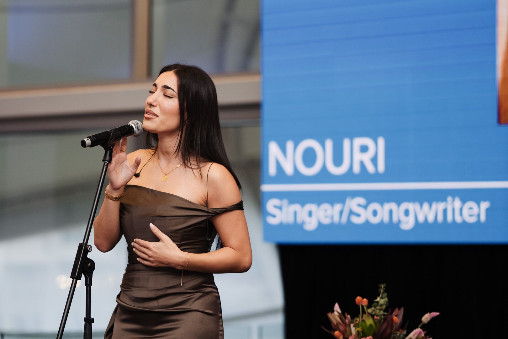 NOURI stands on a stage wearing a copper-coloured dress and sings into a microphone. Behind her is a blue banner with her name
