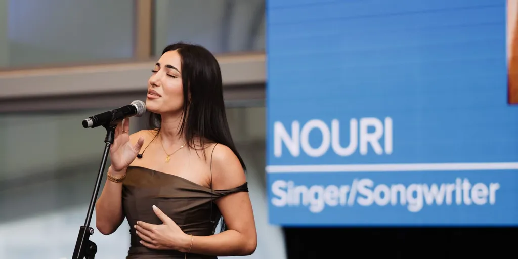 NOURI stands on a stage wearing a copper-coloured dress and sings into a microphone. Behind her is a blue banner with her name