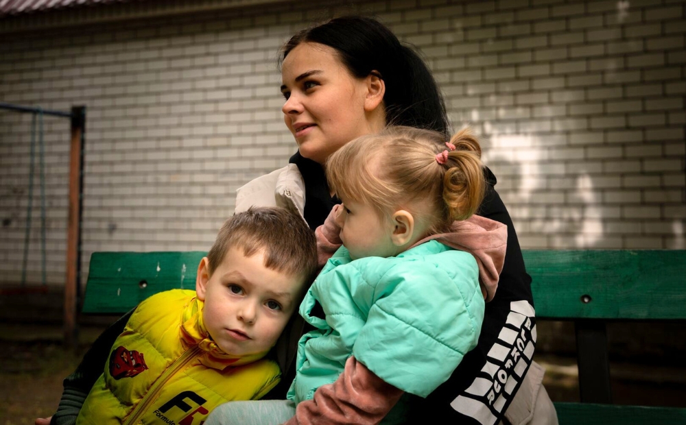 A young woman sits on a bench outdoors, holding her two children; her son wears a yellow jacket and her daughter wears a mint green jacket.
