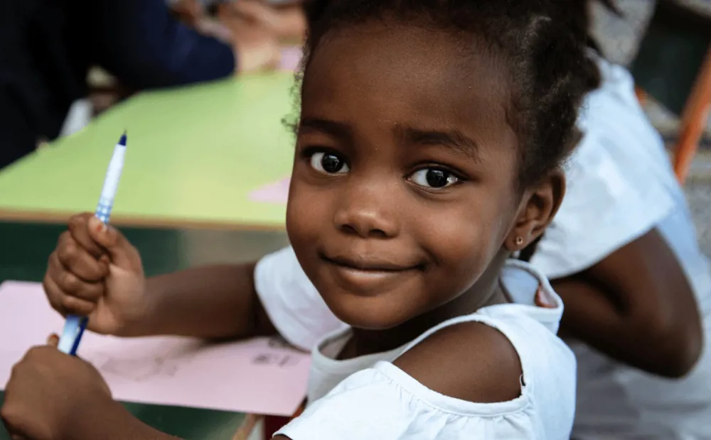 An eight-year-old Tunisian girl sits at a school desk holding a pen and smiling at the camera