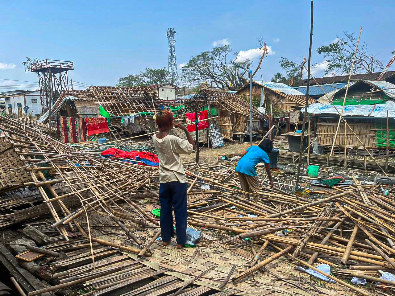 Two refugees in the middle of destroyed shelters in Rakhine state. Shelters at Kyauktaw City Hall IDP site in Rakhine State (Central) which have been destroyed during cyclone Mocha ; Humanitarian needs in Rakhine State and North West Myanmar were already dire before the cyclone hit, with approximately six million people already in need of relief aid