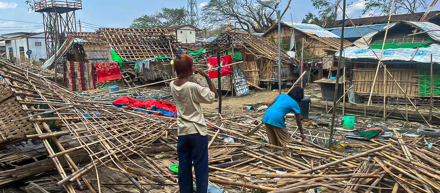 Two refugees in the middle of destroyed shelters in Rakhine state. Shelters at Kyauktaw City Hall IDP site in Rakhine State (Central) which have been destroyed during cyclone Mocha ; Humanitarian needs in Rakhine State and North West Myanmar were already dire before the cyclone hit, with approximately six million people already in need of relief aid