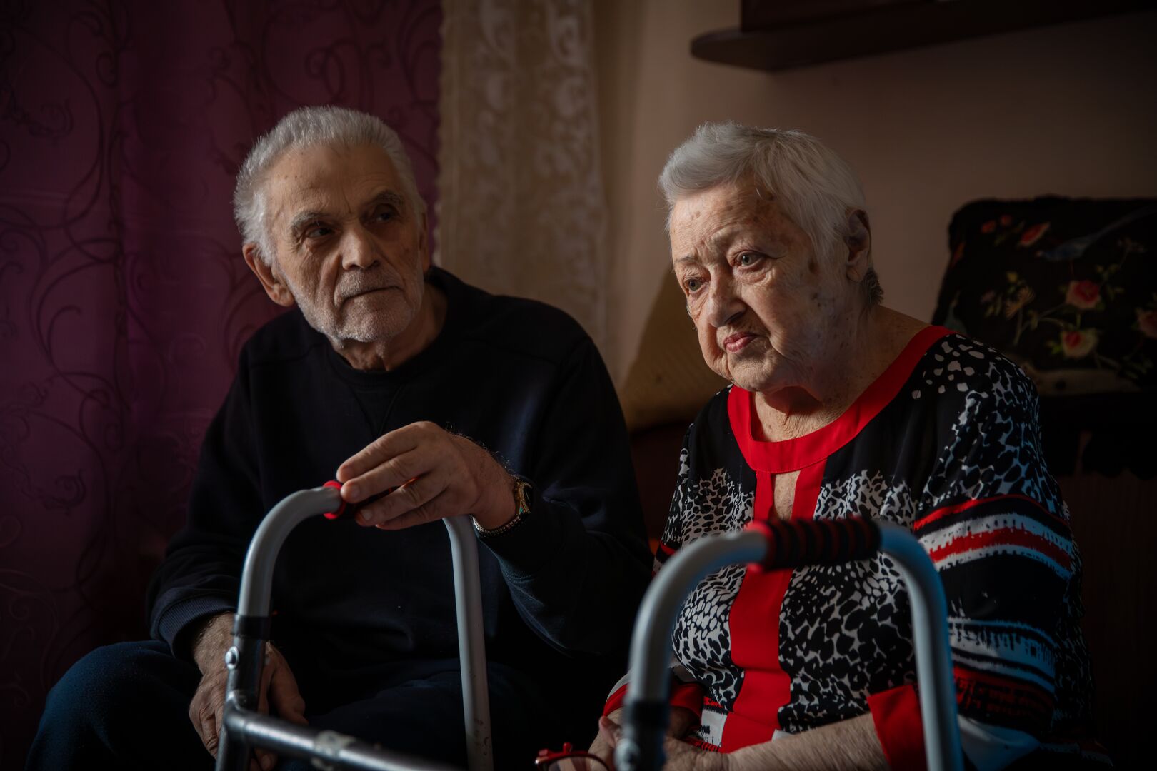 An elderly couple sits together indoors; Valentyna wears a red and black dress and uses a walker, Valentin sits beside her in a dark sweater.