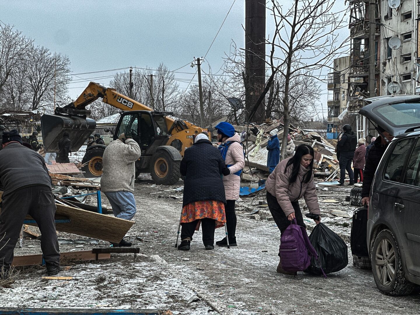Several people, including women and children, gather outdoors among rubble and damaged buildings; a yellow excavator is clearing debris in the background.