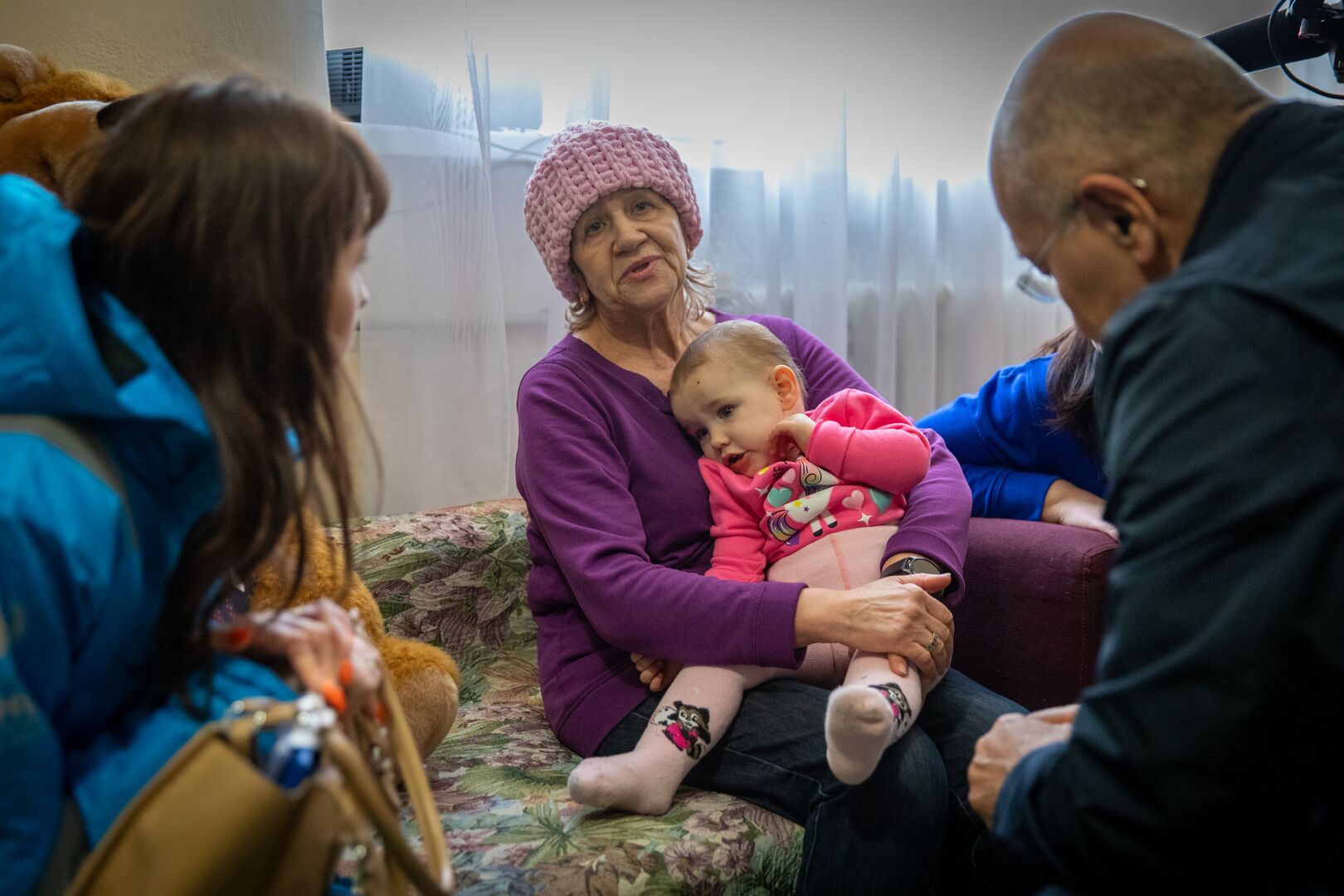 An older woman in a pink knit hat sits on a couch holding a toddler girl in a pink outfit; UNHCR representatives are seated nearby, and stuffed toys are visible in the background.