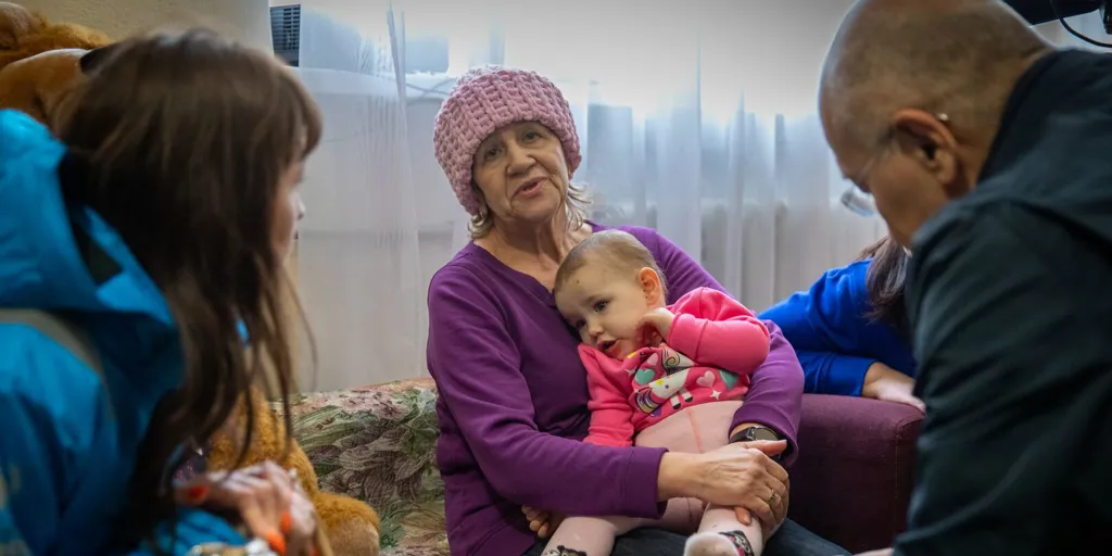 An older woman in a pink knit hat sits on a couch holding a toddler girl in a pink outfit; UNHCR representatives are seated nearby, and stuffed toys are visible in the background.