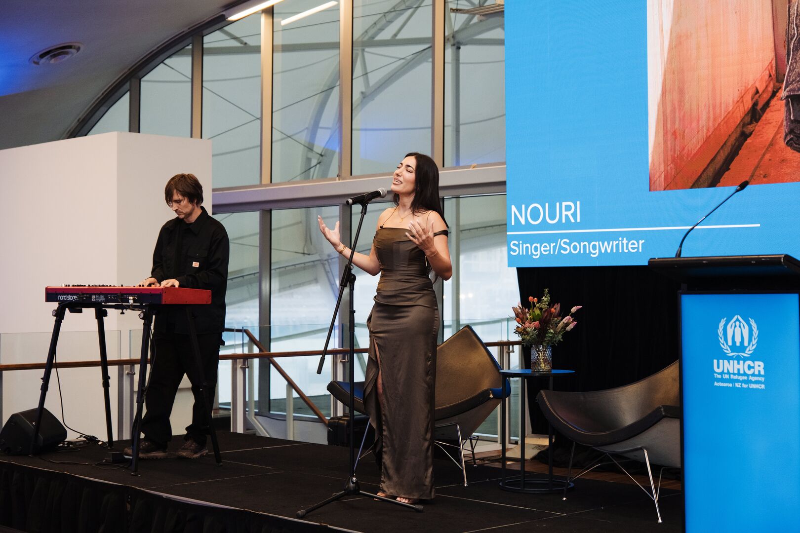 NOURI stands on a stage singing into a microphone. Next to her is a keyboardist and his instrument on a stand. Behind them is a blue banner with her name