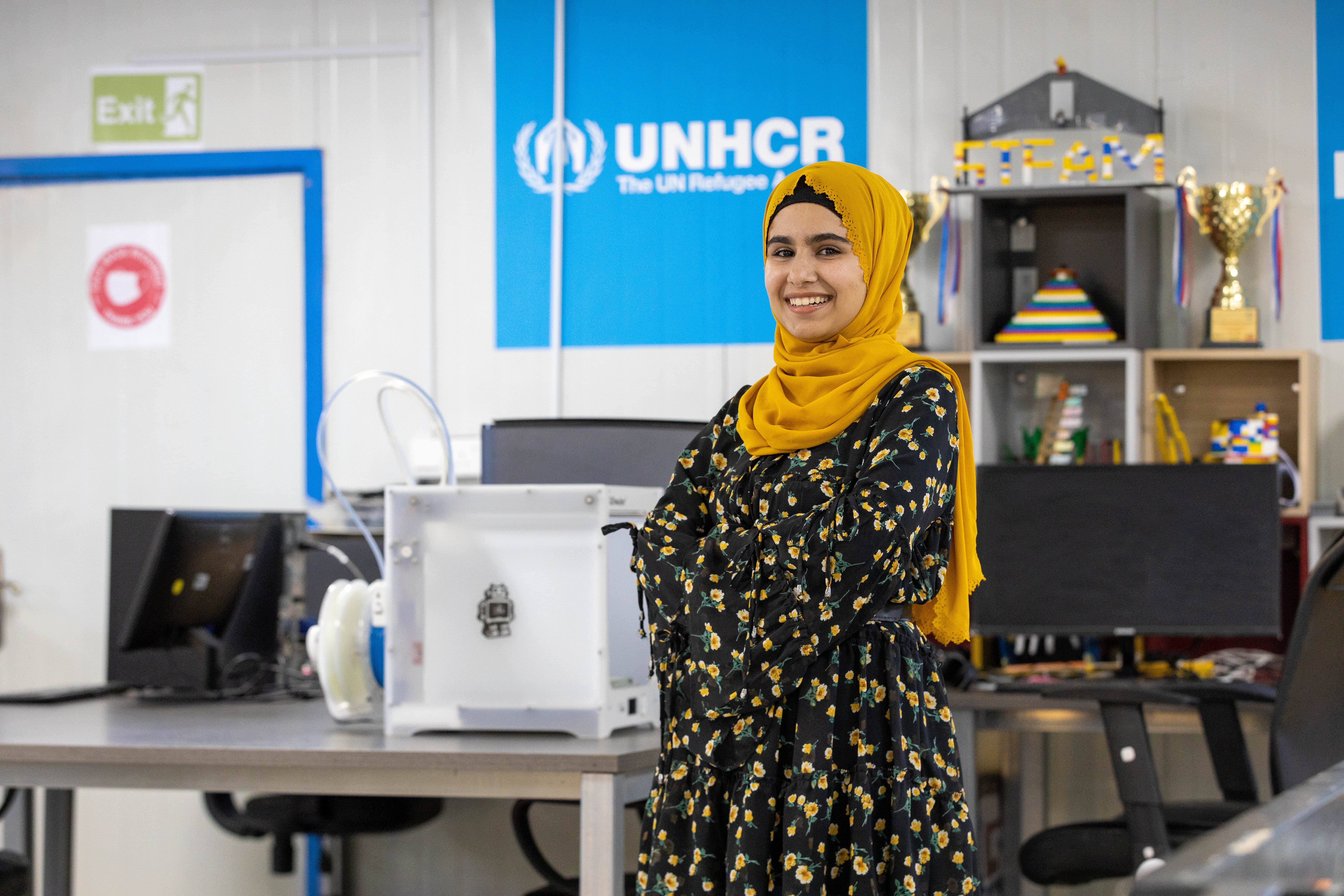 Woman smiles at camera with UNHCR sign behind her. UNHCR’s Innovation Lab promotes STEM for refugee girls. 